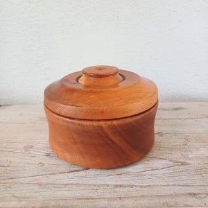 May include: A round, light brown wooden container with a matching lid. The container features a smooth, polished finish that highlights the natural grain patterns of the wood. The lid has a small, raised circular knob at the very centre.