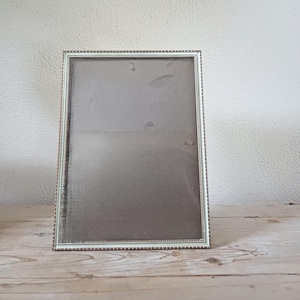 May include: A rectangular silver picture frame with a beaded border. The frame has a glass front and a grey backing. The frame is displayed on a wooden surface against a white wall.