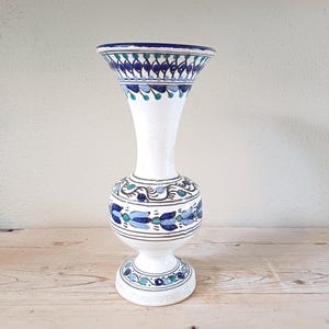May include: White ceramic vase with blue and green floral patterns. The vase has a wide mouth and a narrow neck.