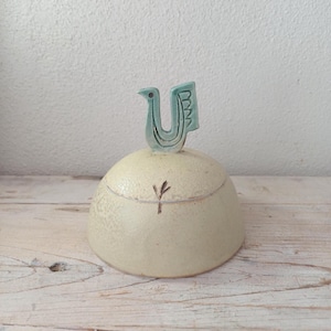 May include: A ceramic lidded jar with a speckled cream-coloured finish and a dome-shaped lid. The lid features a stylised, light teal-coloured bird-shaped handle with incised line details and a small, dark brown hand-drawn plant motif on the side.