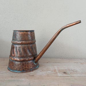 May include: A hammered copper vessel with a wide, cylindrical base and a long, slender, angled spout. The exterior surface features a textured, dimpled finish with horizontal bands encircling the body, showing a warm, aged metallic patina.