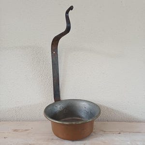 May include: An antique metal ladle with a copper bowl and a long, curved handle. The handle is dark and has a hook at the end for hanging. The bowl is round and shows signs of wear, indicating age and use. The ladle is likely for cooking.