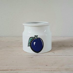 May include: White ceramic jar with a blue plum and green leaf design. The jar is approximately 10 cm tall.