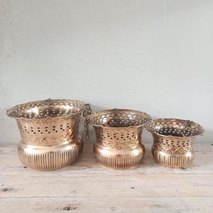 May include: A set of three graduated brass planters with a metallic gold finish. Each planter features a scalloped rim, a decorative band of cut-out heart and arch patterns, and a vertically ribbed base. Metal chains are attached to the rims.