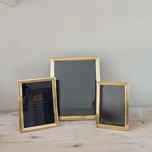 May include: Three rectangular solid brass picture frames with a polished gold-tone finish. The left frame contains a black insert with the text SOLID BRASS 13x18 cm, while the centre and right frames are empty with dark interiors.