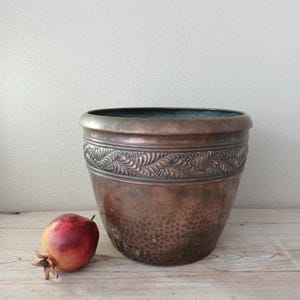May include: A round copper planter with a hammered texture on the lower body and a decorative embossed band featuring a repeating leaf pattern around the upper rim. A single red and yellow pomegranate sits to the left of the metal vessel.