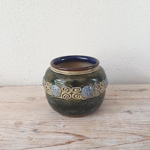 May include: A round ceramic vase with a dark green base and a blue interior. The vase features a decorative band of blue and white swirl patterns around the middle. The rim is a light brown colour.