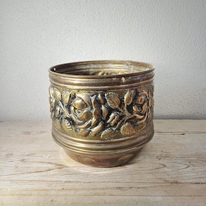 May include: A cylindrical brass planter featuring a prominent embossed floral design of roses and leaves wrapping around the exterior. The metal has a warm, aged patina with dark recessed areas that highlight the intricate relief of the flowers.