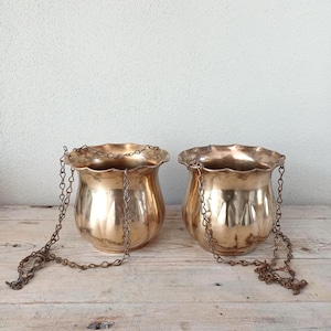 May include: Two matching hanging planters crafted from polished brass, each featuring a rounded, fluted body with a scalloped rim. These metallic vessels are suspended by thin, interconnected metal chains attached to the upper edges of the pots.