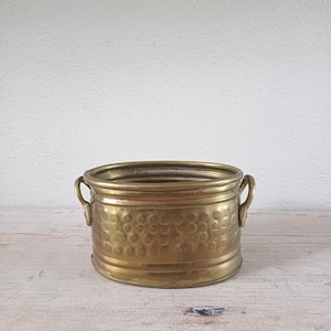 May include: A vintage brass planter with a hammered texture and two small handles. The round planter is a warm gold colour and sits on a light-coloured wooden surface. The planter is empty and ready for use.