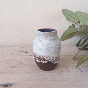 May include: A small, white ceramic vase with a brown rim and a speckled glaze. The vase is sitting on a wooden surface.