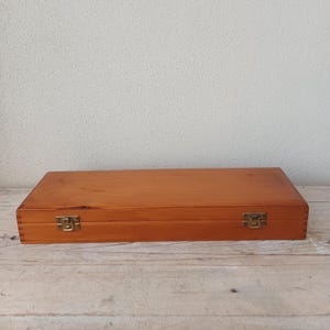 May include: A rectangular wooden storage box with a smooth, light brown finish. The box features visible finger joints at the corners and is secured by two small, metallic brass-coloured latches on the front edge, keeping the lid firmly closed.