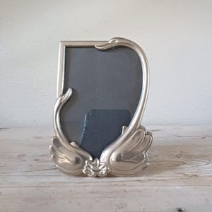 May include: Silver-coloured metal picture frame with a swan design. The frame features two swans, their necks forming the sides of the frame, with a flower detail at the base. The frame holds a dark grey insert.