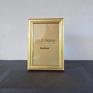May include: A rectangular, gold-coloured solid brass photo frame. The frame is hand polished and lacquer coated. The frame is 4" x 6" (10cm x 15cm) and has decorative detailing around the edges. The text "SOLID BRASS" and "Bowon" are visible.