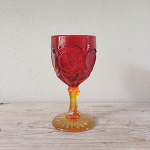 May include: A vintage red glass goblet with a starburst design on the base. The goblet features a vibrant red bowl with a decorative pattern and a yellow and orange stem. The glass is likely used for serving beverages.