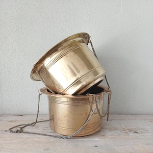 May include: Two stacked brass planters with a polished metallic finish. Each cylindrical container features a wide rim and is equipped with thin metal chains attached to the sides for hanging, showcasing a smooth surface with subtle variations in tone.