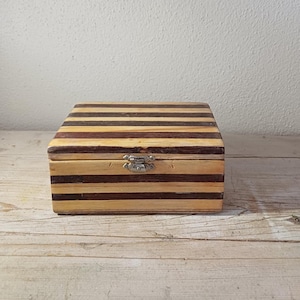 May include: A rectangular wooden box featuring alternating horizontal stripes of light tan and dark brown wood. The box has a hinged lid and a small, silver-coloured metal latch centred on the front edge to secure the closure of the container.
