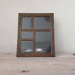 May include: A rectangular shadow box with a decorative gold-coloured frame and multiple glass compartments. The frame features an ornate, swirling pattern. The box is displayed on a wooden surface against a white wall.