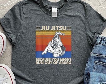 May include: A dark gray t-shirt with a vintage-style graphic of a jiu-jitsu match. The graphic has a sunset background with the text "Jiu Jitsu" and "Because you might run out of ammo".