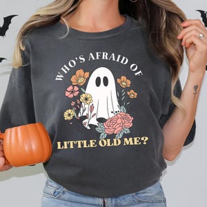 May include: Dark gray t-shirt with the text "WHO'S AFRAID OF LITTLE OLD ME?" featuring a ghost and floral design. An orange pumpkin-shaped mug is held in the hand. Black bat decorations are on the wall.