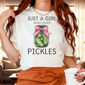 Könnte beinhalten: Cremefarbenes T-Shirt mit dem Text "JUST A GIRL WHO LOVES PICKLES" und einer Grafik eines Einmachglases mit rosa Schleife. Die Person trägt orangefarbene Shorts und hält einen weißen Becher.