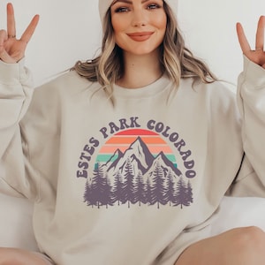 May include: A beige sweatshirt with a vintage-style graphic of mountains, a sunset, and the text "Estes Park Colorado".