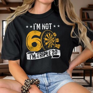 Könnte beinhalten: Schwarzes T-Shirt mit dem Text "I'M NOT 60 I'M TRIPLE 20" in Gelb und Weiß, mit einem Dartscheiben-Design. Das Shirt wird mit Jeansshorts und weißen Turnschuhen kombiniert.