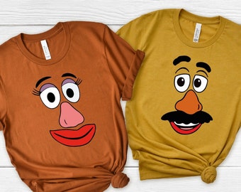 May include: Two brown and mustard yellow t-shirts with a cartoon image of Mr. and Mrs. Potato Head. The shirts are folded and laid out on a white wooden surface.