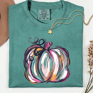 Könnte beinhalten: Türkisfarbenes Comfort Colors T-Shirt mit einem farbenfrohen, abstrakten Kürbisdesign. Der Kürbis hat rosa, weiße, orangefarbene und türkisfarbene Striche. Eine goldene Halskette mit Sternanhänger befindet sich auf dem Shirt. Größe XL-TG-EG.