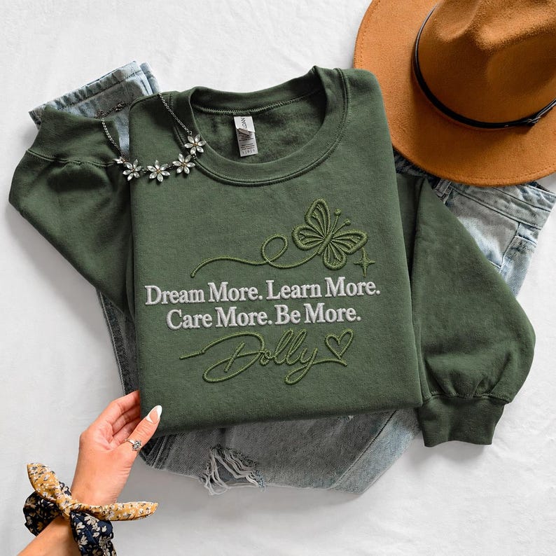 A green sweatshirt with a butterfly design and the text "Dream More. Learn More. Care More. Be More." is displayed on a bed, along with a hat and a pair of jeans. A hand is visible in the foreground, holding a bracelet.