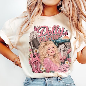 May include: A cream-colored t-shirt featuring a graphic print of Dolly Parton with a guitar, pink cowboy boots, butterflies, and flowers. The text reads Dolly Parton, Smoky Mountain Girl, and Find out who you are and do it on purpose.