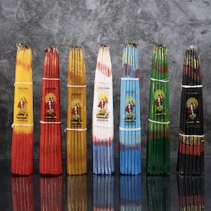 Pode incluir: Um conjunto de dez velas, cada uma embrulhada em uma cor diferente, com um rótulo dourado que diz "Terra Santa Jerusalém". As velas estão dispostas em uma fileira sobre uma superfície cinza.