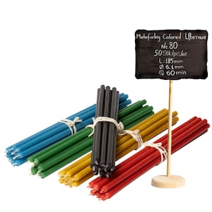 Puede incluir: Un conjunto de 50 velas de colores, agrupadas en cinco colores diferentes: azul, verde, amarillo, negro y rojo. Las velas tienen un diámetro de 6,1 mm y una longitud de 185 mm. Un letrero de pizarra dice: "Mehrfarbig Colored Цветные No 80 50 Stk/pcs/шт. L:185mm Ø 6.1 mm 60 min".