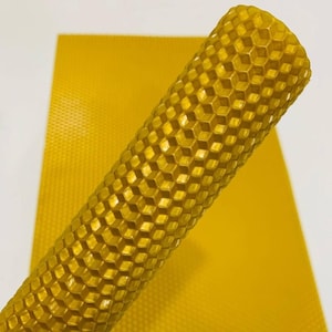 Può includere: Primo piano di fogli di cera d'api giallo dorato. Un foglio è arrotolato, mostrando un motivo a nido d'ape. L'altro foglio è piatto, mostrando lo stesso design esagonale. I fogli hanno un colore e una consistenza caldi e naturali.