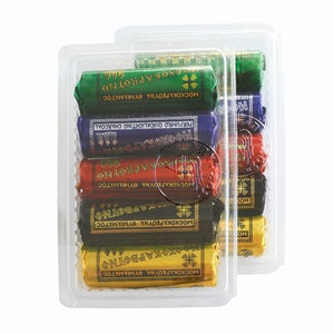 May include: Two clear plastic containers filled with stacks of colourful incense sticks. Each stick is wrapped in gold foil and labelled with Greek text. The text reads "MOSCHOKARBOUNA THYMIAMATOS" which translates to "Perfumed Charcoal Incense".
