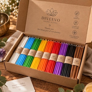 Peut inclure: Une boîte en carton contenant des rangées de bougies fines en cire d'abeille colorées, incluant blanc, bleu, vert, jaune, orange, rouge, violet, noir et marron. Le couvercle porte le texte DIVEEVO NATURAL RITUALS et une icône de lotus.