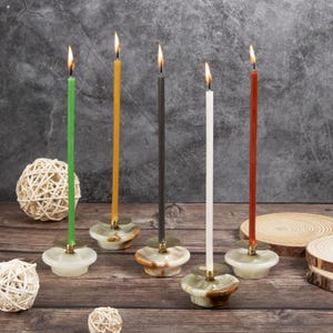 Peut inclure: Cinq bougies allumées de différentes couleurs, vert, jaune, noir, blanc et rouge, sont placées sur des bougeoirs en marbre décoratifs. Les bougeoirs sont sur une surface en bois.