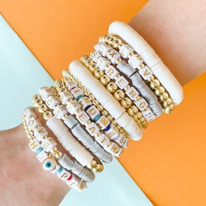 May include: A stack of bracelets with white beads and gold beads with words like "LOVE", "HOPE", "KINDNESS", "STRENGTH", and "LUV" spelled out on them.
