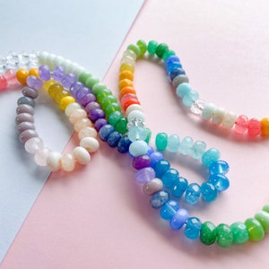 May include: A colorful beaded necklace with a rainbow of colors, including pink, orange, yellow, green, blue, purple, and white. The beads are made of various materials, including glass, stone, and wood.