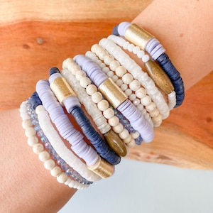 May include: A stack of bracelets in shades of white, lavender, and navy blue. The bracelets are made of various materials, including beads, stones, and metal accents.