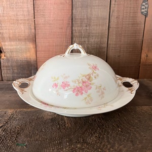 May include: A white porcelain butter dish with a floral design. The dish has a domed lid with a handle and a base with two handles. The floral design features pink flowers and green leaves.