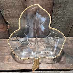 May include: Clear glass leaf-shaped serving dish with a gold-colored trim and stem. The dish features detailed vein patterns and is designed for serving snacks or small items. The leaf dish is a decorative piece.