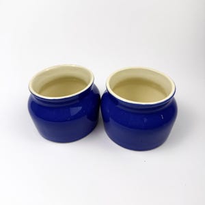 May include: Two small, round ceramic pots with a glossy, deep blue exterior finish and a smooth, cream-colored interior. Each vessel features a wide, open top and a slightly tapered base, presenting a simple, functional design for storage.