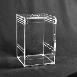 May include: A clear, rectangular acrylic box with a hinged door and small ventilation holes. The box has rows of small, evenly spaced dots along the edges. The box is designed to hold small items.