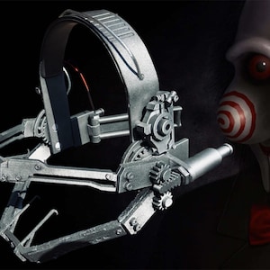 May include: A silver metal trap with a gear system and a long, thin metal arm. The trap is designed to be worn on the head and has a strap that goes around the back of the head. The trap is a prop from the horror movie Saw.
