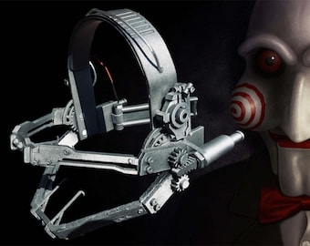 Könnte beinhalten: Eine silberne Metallfalle mit einem Getriebesystem und einem langen, dünnen Metallarm. Die Falle ist so konzipiert, dass sie auf dem Kopf getragen werden kann und ein Band hat, das um den Hinterkopf geht. Die Falle ist eine Requisite aus dem Horrorfilm Saw.