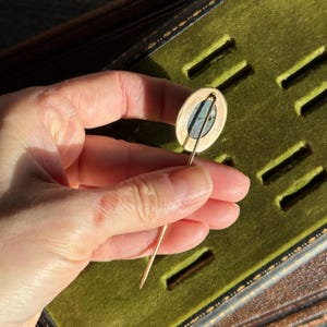 May include: A gold-toned stick pin with an oval head. The head features a central, possibly stone, element with blue and green hues, set within a gold frame. The pin is held against a green velvet surface.
