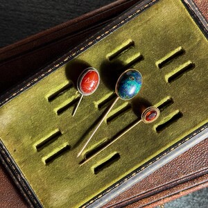 May include: Three antique hat pins with decorative gemstone heads. The pins are displayed in a green velvet-lined box. One pin has a red stone, another a blue and green stone, and the third a red-orange stone.