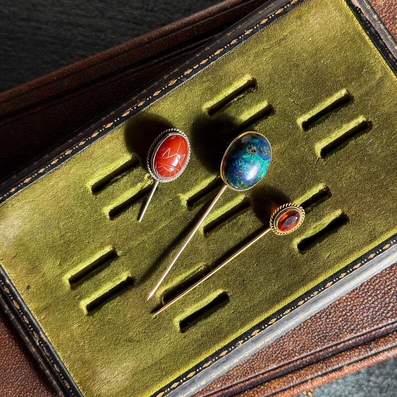 May include: Three antique hat pins with decorative gemstone heads. The pins are displayed in a green velvet-lined box. One pin has a red stone, another a blue and green stone, and the third a red-orange stone.