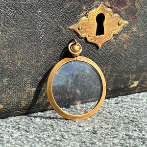 May include: A circular gold-toned pendant featuring a clear glass center and a small, rounded bail at the top. The pendant has a smooth, polished metal frame surrounding the transparent circular pane, designed for use as a locket or decorative charm.